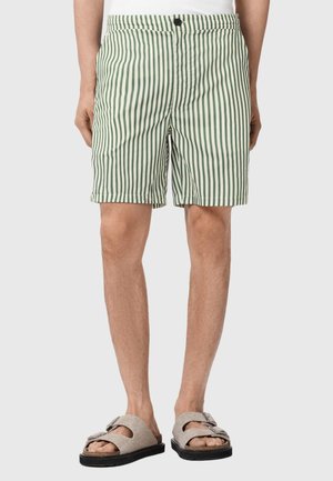 Mand iført grønne og hvide vertikalt stripede shorts og beige sandaler med sorte såler, stående mod en ensartet lysegrå baggrund.