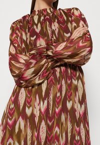 Lång, flödande klänning med hög krage, med en brun bas och djärva abstractmönster i rosa, vitt och beige. Texturerade puffärmar.