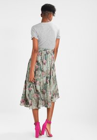 Jupe midi florale avec un design fluide en vert, ornée de fleurs roses et crème, associée à un haut gris à manches courtes et des escarpins roses.