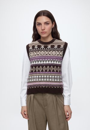 Femme portant un gilet en maille marron à motifs sans manches par-dessus une chemise blanche à manches longues et un pantalon plissé beige, debout devant un fond uni.