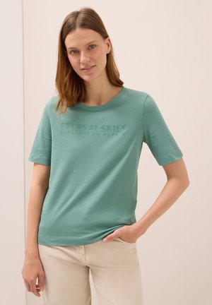 Femme avec des cheveux raides châtain clair portant un t-shirt vert avec le texte "CIEUX ÉTERNELS" et un pantalon beige, se tenant avec une main dans la poche.
