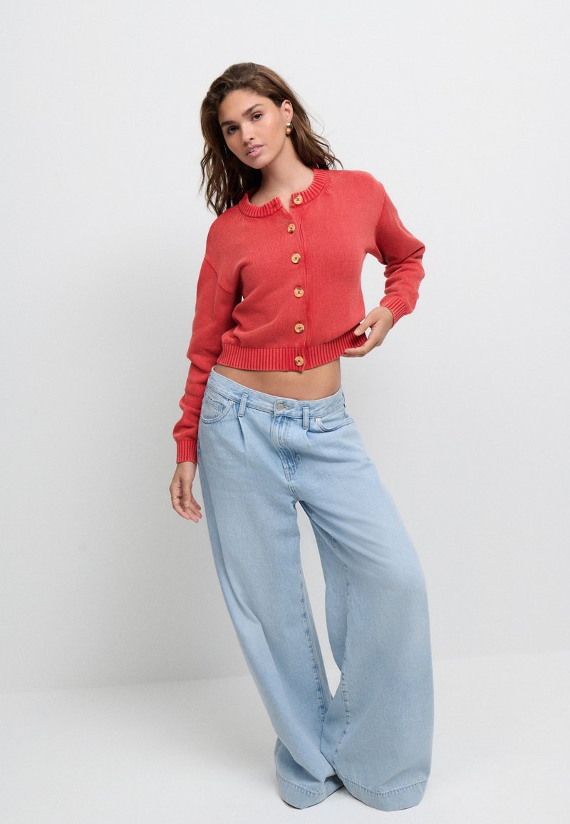 Coral knitted cardigan with a ribbed collar and six wooden buttons. Paired with light blue wide-leg jeans, featuring a relaxed fit.