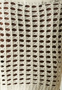 Dzianinowy top na ramiączkach w kolorze kremowym, z ażurowym wzorem z dużymi owalnymi i okrągłymi motywami oraz prążkowanym dekoltem.