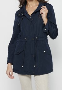 Femme portant une veste à capuche bleu marine avec fermeture éclair avant, boutons-pression, cordons, poches à rabat, et un pantalon beige, vue du cou jusqu'au milieu des cuisses.