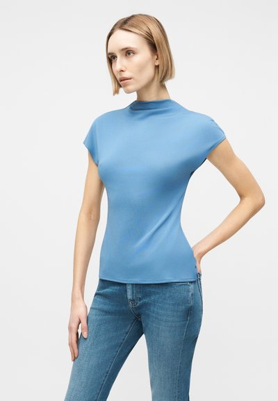 Γυναίκα με κοντά ξανθά μαλλιά που φοράει στενό ανοιχτό μπλε κοντομάνικο πάνω μέρος και μπλε τζιν, όρθια με το ένα χέρι στη μέση.