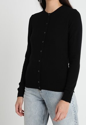 Zwarte cardigan van zachte stof, met een ronde halslijn, lange mouwen en een knoopsluiting aan de voorkant. Gedragen met lichtblauwe jeans.