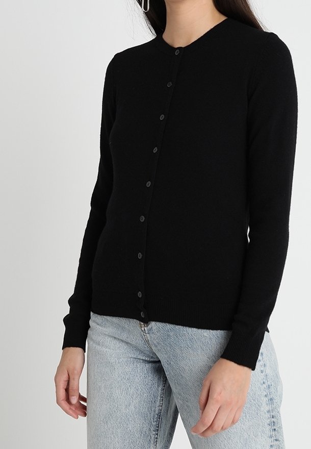 Zwarte cardigan van zachte stof, met een ronde halslijn, lange mouwen en een knoopsluiting aan de voorkant. Gedragen met lichtblauwe jeans.