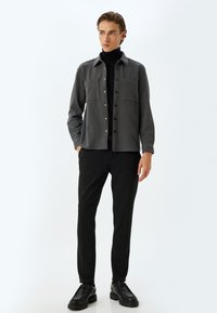 Grå skjorte med knapper og to brystlommer, båret over en sort turtleneck. Kombineret med sorte bukser og sorte snørebukser, på en neutral baggrund.