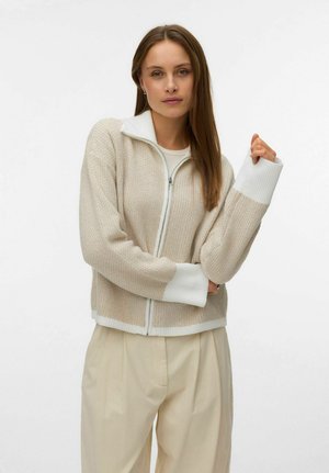 Kvinne med langt brunt hår iført en beige genser med glidelås, hvite mansjetter og krage, og løse beige bukser, poserende mot en hvit bakgrunn.