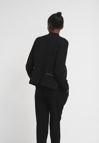 Veste noire à coupe décontractée, conçue avec un col montant, un détail de poche arrière unique et un tissu lisse, accompagnée d'un pantalon assorti.