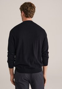 Pull en tricot noir à manches longues, avec poignets et ourlet côtelés, texture lisse et col rond classique, porté avec un pantalon foncé.
