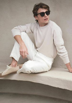 Jonge man met zonnebril, beige trui, witte broek en beige sneakers, casual zittend tegen een lichtgrijze gebogen muur.