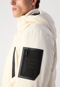 Helle Creme Pufferjacke mit Kapuze, ausgestattet mit einer mattschwarzen Reißverschlusstasche am Ärmel und einem dezenten Logo auf einem quadratischen Patch.