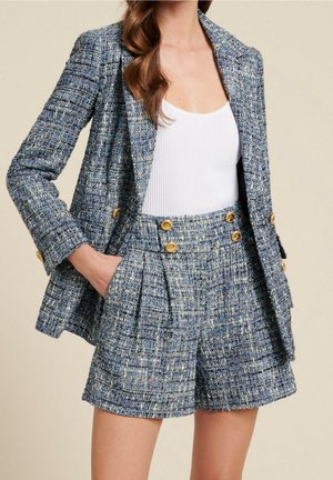 Femme portant un blazer en tweed bleu et un short plissé taille haute assorti avec des boutons dorés, associé à un débardeur blanc côtelé.