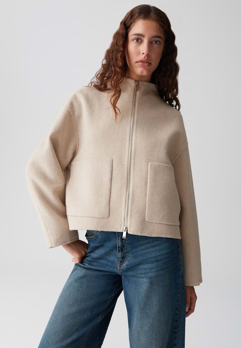 Giacca corta beige realizzata in tessuto morbido, con colletto alto, zip frontale e due ampie tasche quadrate. Abbinata a jeans blu.
