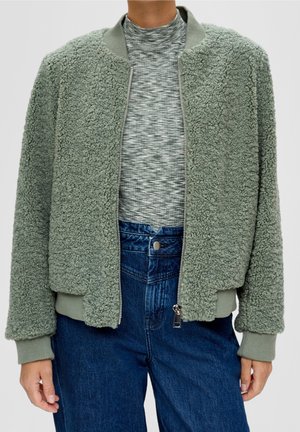 Personne portant une veste sherpa en polaire verte, ouverte, sur un col roulé gris à motifs et un jean bleu taille haute, les bras détendus le long du corps.