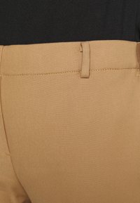 Pantalons beige en tissu tissé, présentant une texture lisse, une coupe droite et un discret détail de couture jaune à la taille.