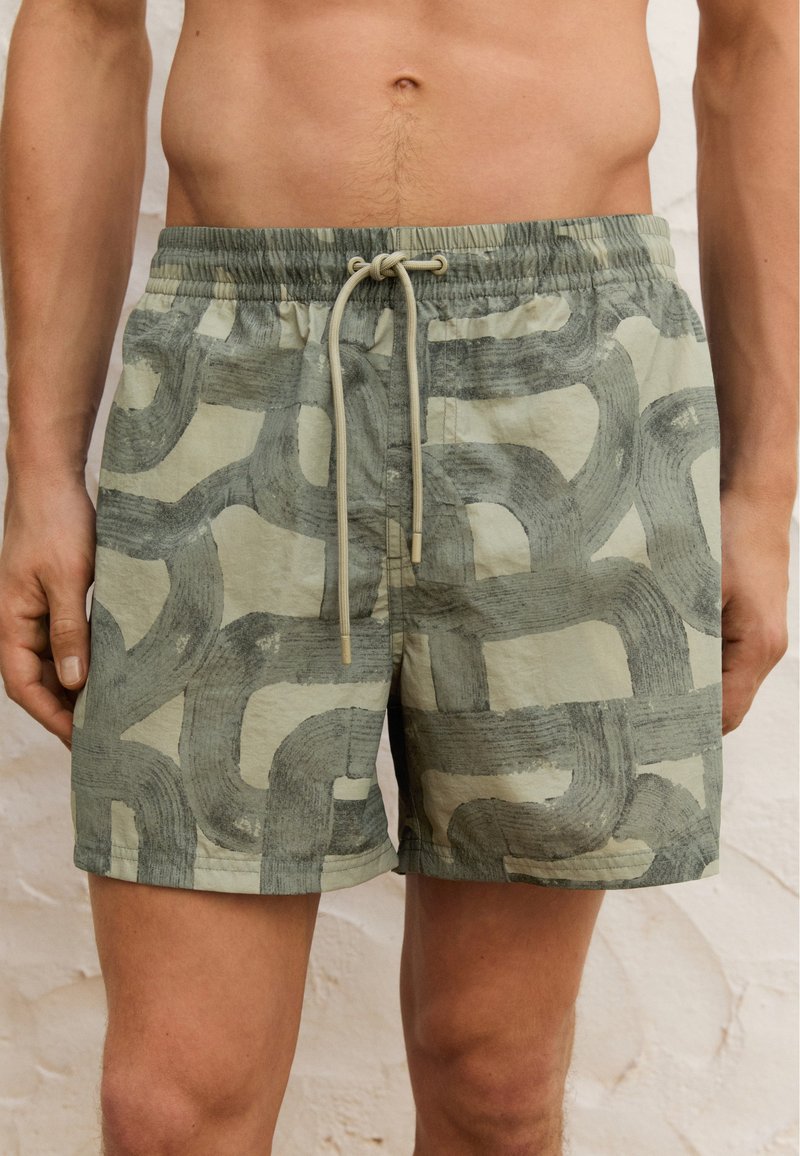 Schwimmshorts aus leicht grünem Stoff mit abstraktem dunkelgrünen Pinselstrichmuster; elastischer Bund mit verstellbarem Kordelzug; mittellange Beine.