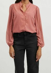 Rosa blus med zickzackmönster, långa puffärmar och hög krage, med knappar framtill. Matchad med svarta jeans.