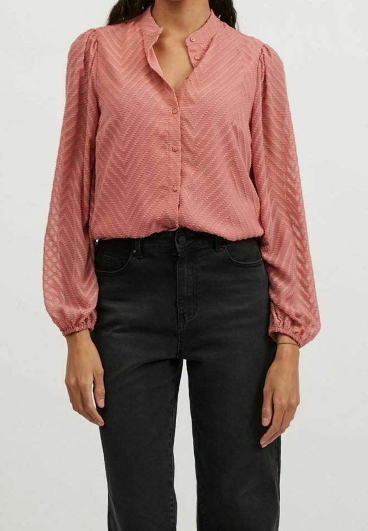 Rosa blus med zickzackmönster, långa puffärmar och hög krage, med knappar framtill. Matchad med svarta jeans.