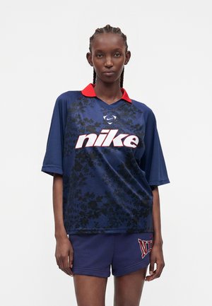 Γυναίκα φοράει μπλε αθλητικό μπλουζάκι Nike με κόκκινο γιακά και ταιριαστά μπλε σορτς με το λογότυπο της Nike, στέκεται μπροστά σε απλό ανοιχτό φόντο.