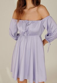 Vestido lilac fuera de los hombros con mangas largas abullonadas, cintura elástica y detalle de cordón; hecho de una tela ligera con una textura suave.