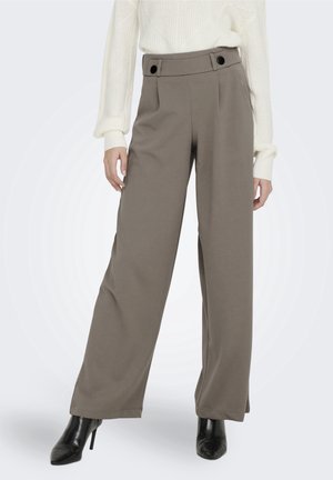 Frau trägt hoch taillierte, weit geschnittene Hose in Taupe mit schwarzen Knopfdetails an der Taille, einen weißen Strickpullover und schwarze spitze, hochhackige Stiefel.