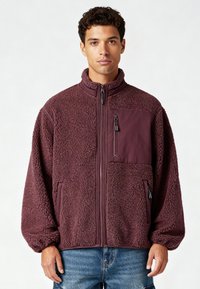 Giacca in pile bordeaux con collo alto, chiusura frontale con zip e tasca sul petto. Tessuto morbido e strutturato con polsini elastici e tasche laterali.