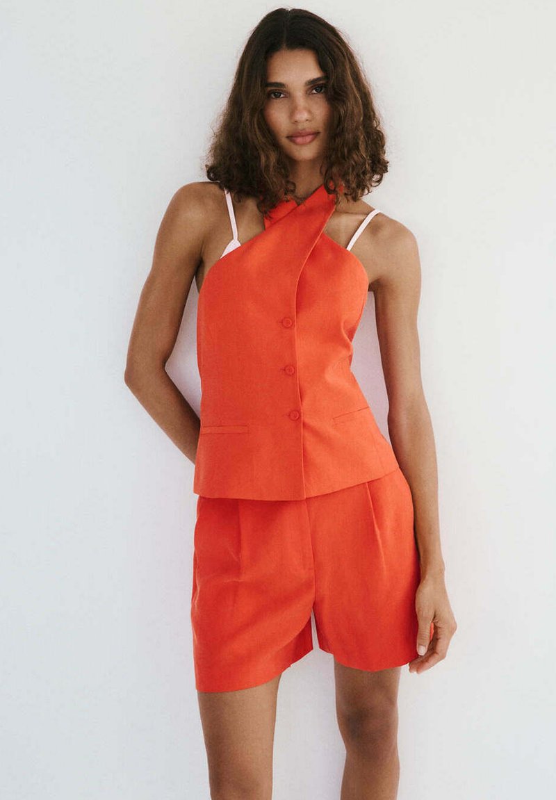 Femme aux cheveux bouclés portant un haut sans manches orange vif avec boutons et un short assorti, debout devant un fond clair uni.