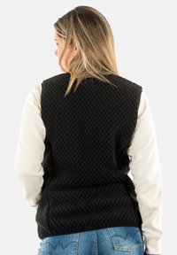 Gilet trapuntato nero con superficie testurizzata, abbinato a una camicia bianca a maniche lunghe, indossato sopra un paio di jeans in denim.