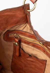 Binnenkant van een bruine leren tas met een grote ritszak, beige voering en een extra kleiner zakje. Voorzien van een messing ring als hardware detail.