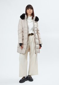 Giacca a vento beige chiaro con colletto in pelliccia nera, caratterizzata da un design trapuntato, cerniera frontale e tasche laterali; indossata sopra un maglione bianco e pantaloni crema.