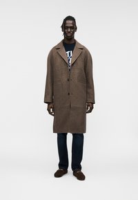 Καφέ παλτό από μαλλί με γιακά με εγκοπή, κλείσιμο με ένα κουμπί και πλαϊνές τσέπες, φορεμένο πάνω από ένα γραφικό πουκάμισο, σε συνδυασμό με σκούρα τζιν και παντόφλες.