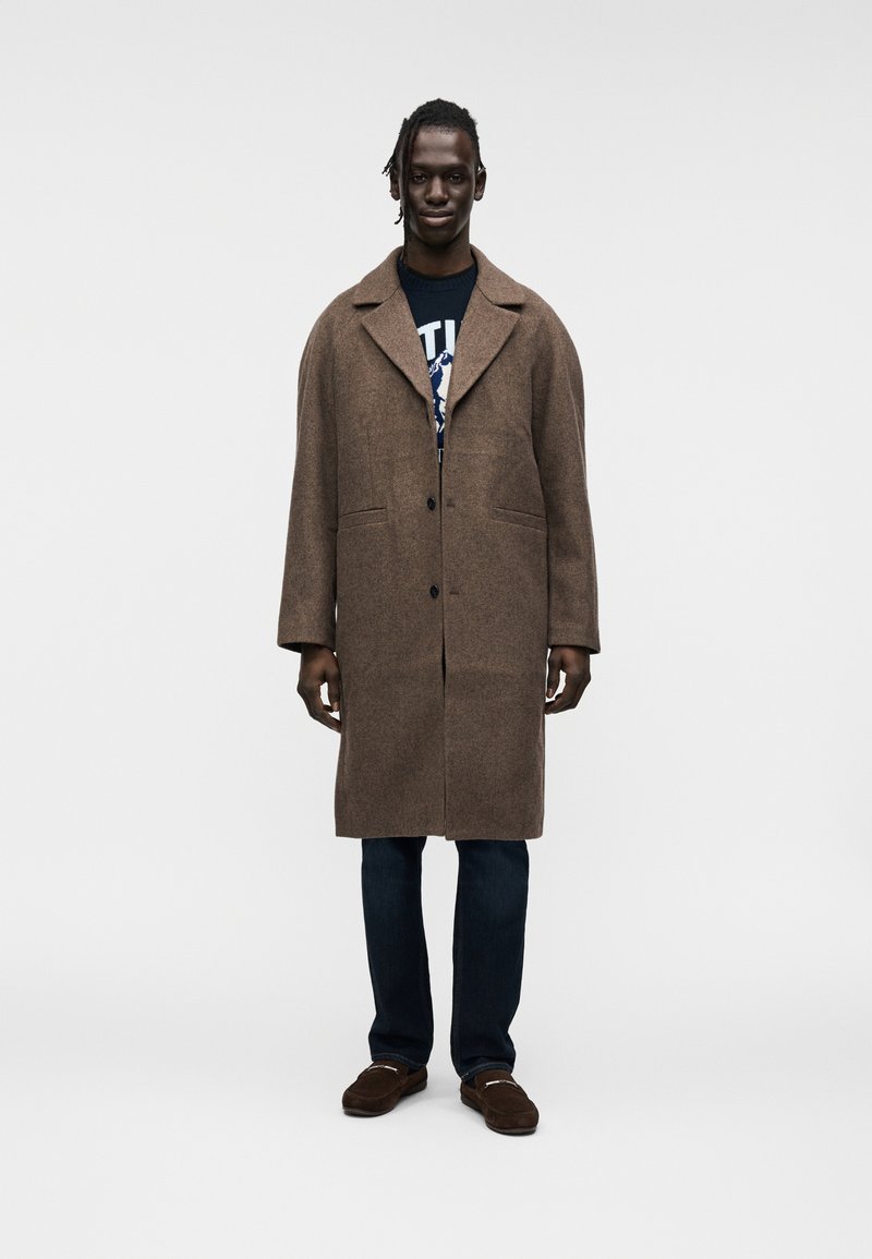 Καφέ παλτό από μαλλί με γιακά με εγκοπή, κλείσιμο με ένα κουμπί και πλαϊνές τσέπες, φορεμένο πάνω από ένα γραφικό πουκάμισο, σε συνδυασμό με σκούρα τζιν και παντόφλες.