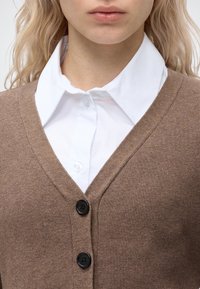 Femme portant une chemise blanche à col sous un cardigan marron boutonné, montrant l'encolure et le haut de la poitrine.