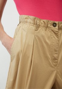 Primo piano di una persona che indossa pantaloni beige a vita alta con un bottone e pieghe, mano in tasca, abbinati a un top in maglia a coste rosa brillante.