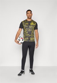 Camisa negra de manga corta con texto gráfico amarillo y un logo circular, combinada con pantalones negros y zapatillas deportivas negras, sosteniendo un balón de fútbol.