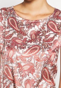 Blusa a maniche corte con scollo rotondo in tessuto leggero, caratterizzata da una stampa paisley in rosso, rosa e marrone con accenti floreali e una texture liscia.