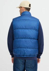 Zilas pūkainais bezmugurs veste ar augstu apkakli, kurai ir izšūti sektori, gluda tekstūra un pieguļošs viduklis. Apvienots ar tumši zilu garām piedurknēm kreklu.