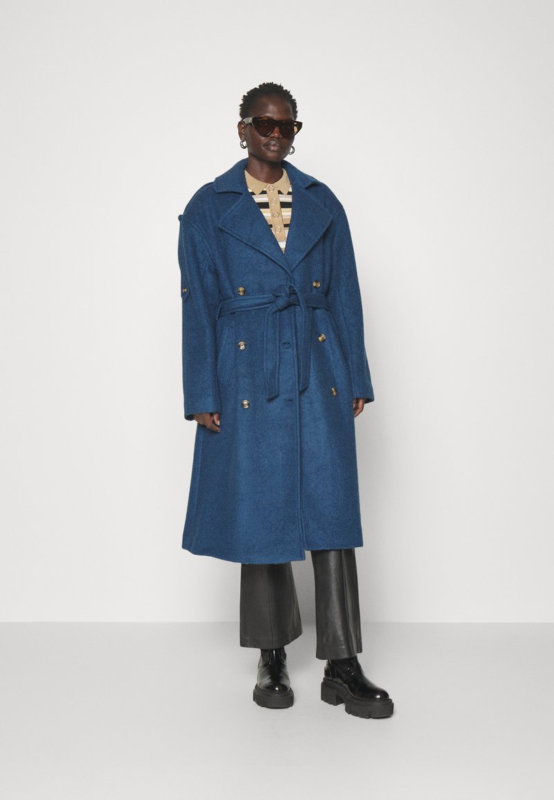 Niebieski płaszcz wykonany z teksturowanej wełny z podwójnym zapięciem, dużymi klapami i paskiem. W zestawie z czarnymi skórzanymi spodniami i masywnymi butami.