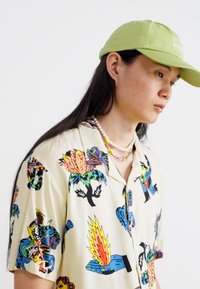 Chemise à manches courtes avec un fond crème, ornée d'imprimés graphiques colorés de mains et de plantes. Association avec une casquette verte et un collier en perles.