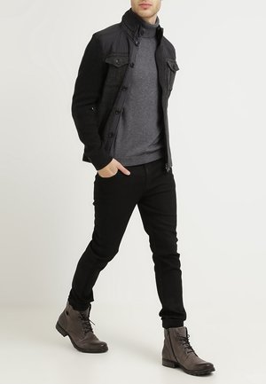 Hombre caminando con una chaqueta negra, suéter de cuello alto gris, pantalones negros y botas marrones con cordones sobre un fondo liso.