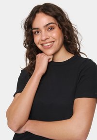 Crna rebrasta majica kratkih rukava s okruglim izrezom. Glatka tekstura tkanine, prilagođen stil, uz opušteni stav modela.
