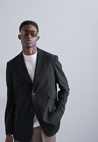 Sacou negru, ajustat, cu două buzunare frontale, purtat peste o cămașă albă. Modelul este accesorizat cu ochelari de soare maro supradimensionați.