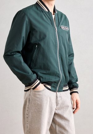 Personne portant une veste bomber vert foncé avec des poignets et un col rayés, un jean gris clair, une main dans la poche, debout devant un fond uni.