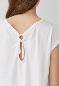 Weißes Kurzarmoberteil mit einem V-förmigen Rückenausschnitt, der mit zwei schmalen weißen Bändern gebunden ist; aus weichem, glattem Baumwollmaterial.