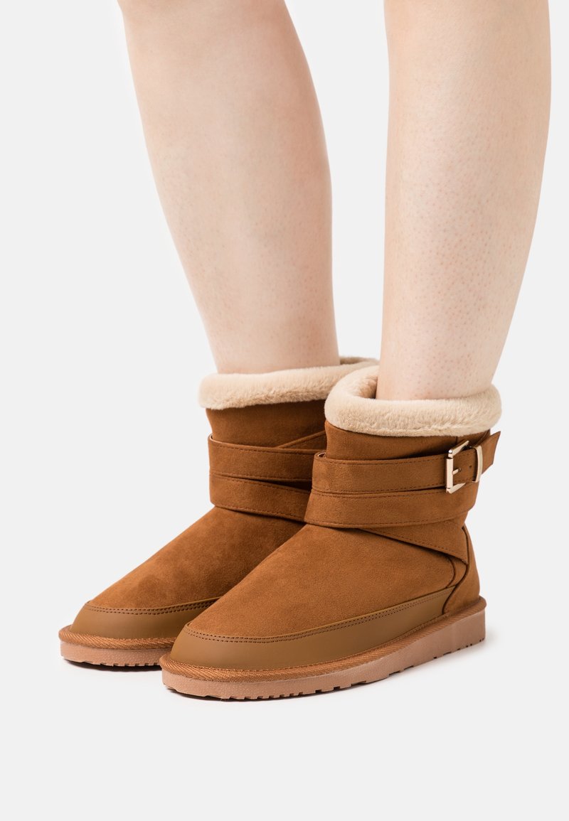 Bottines marron avec doublure en fourrure beige et une bride à boucle, portées sur jambes nues sur un fond clair uni.