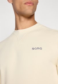 Világosbarna pulóver, puha anyagból, kerek nyakkivágással, sötétkék hímzett "BORG" logóval a mellrészen.