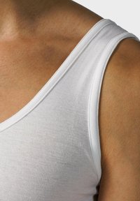 Biały bawełniany top na ramiączkach z szerokimi ramiączkami i okrągłym dekoltem, eksponujący gładką fakturę na opalonej skórze.