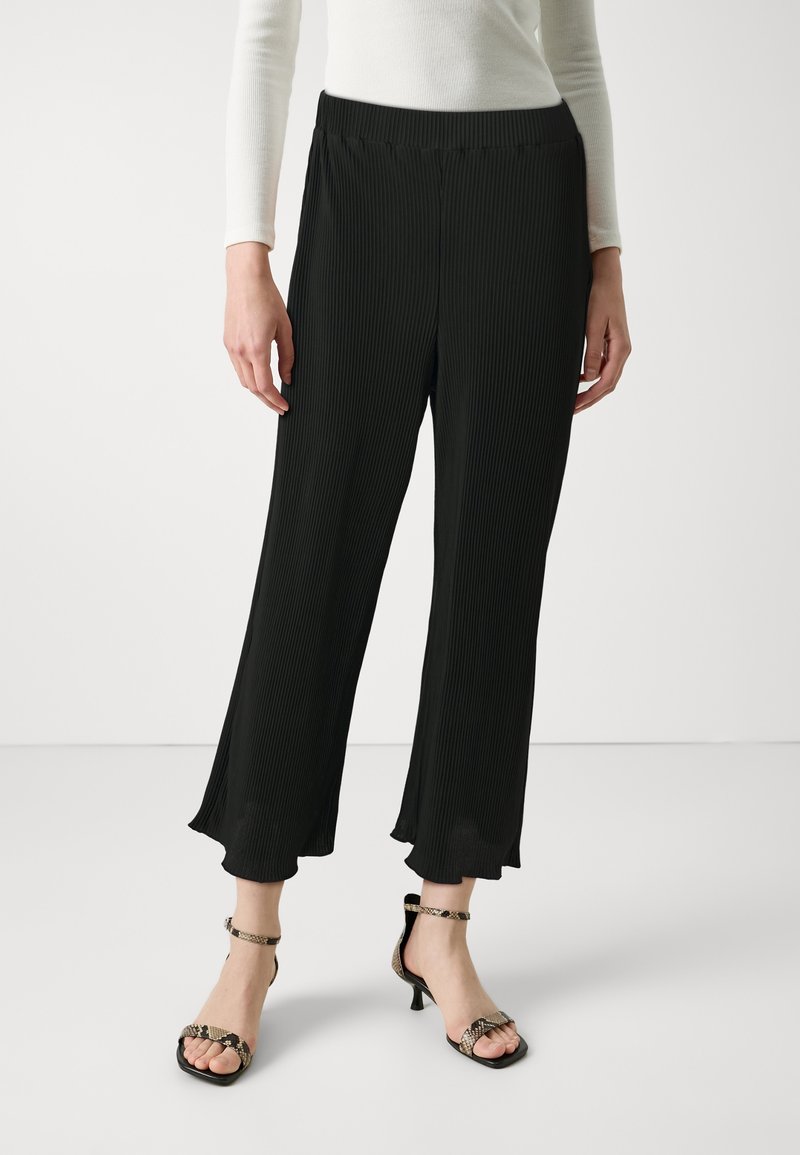 Pantalons larges plissés noirs avec une taille lisse, confectionnés dans un tissu léger et d'une longueur cropped, associés à des sandales à talons avec lanières.